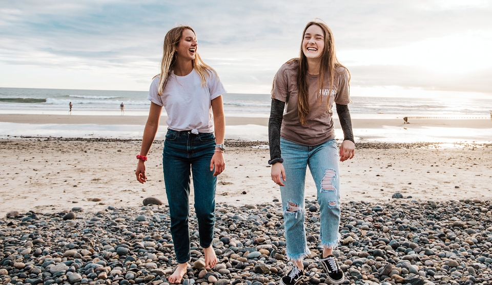 To unge piger går på en strand. Solen skinner i baggrunden og pigerne ser meget glade ud.