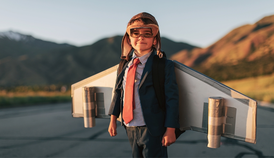 En ung dreng står iført jakke og slips med en gammeldags pilothat på. Han har vinger på ryggen, der minder om dem der er på en flyvemaskine.