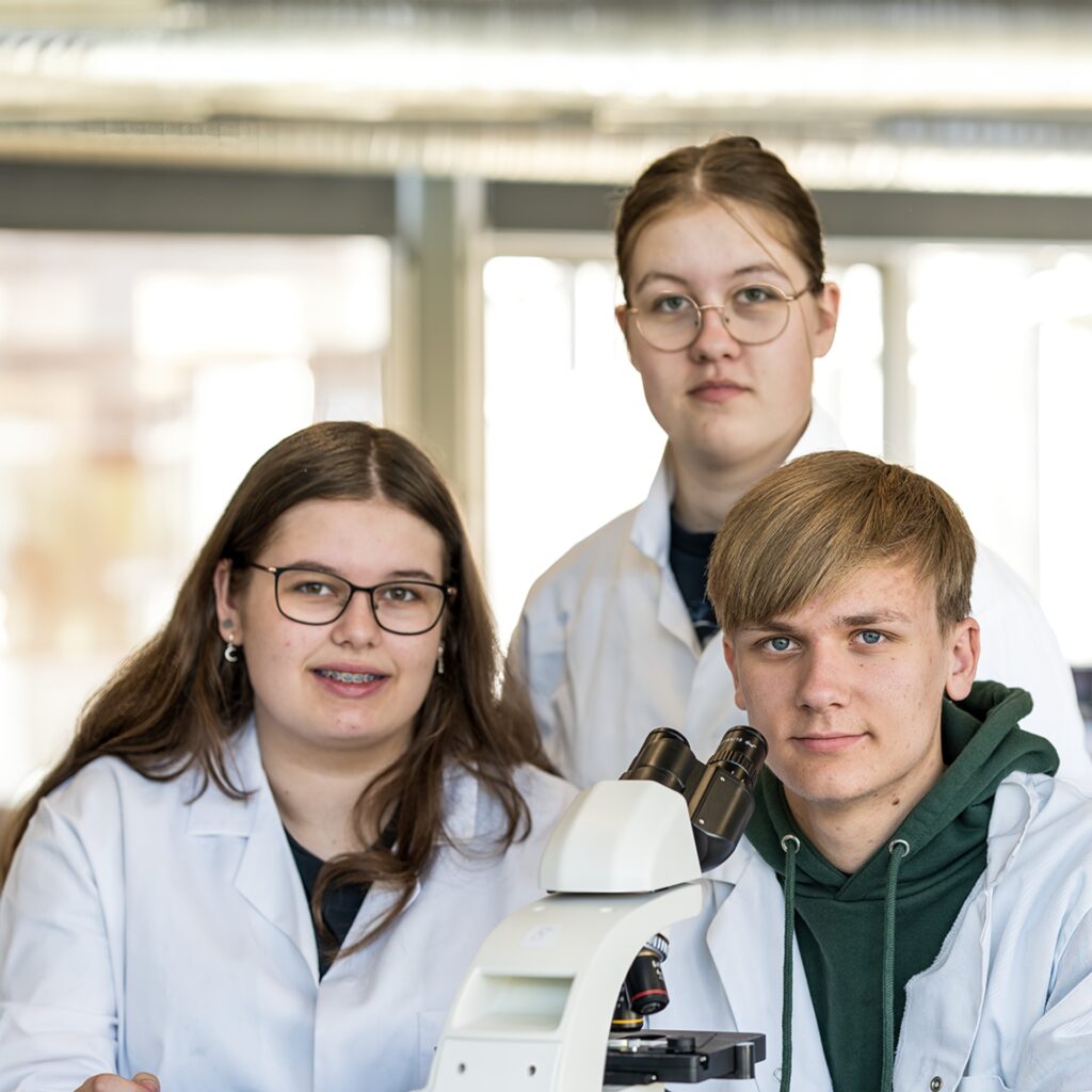 Tre elever fra NEG Kalundborg er iført kitler og sidder ved et mikroskop.