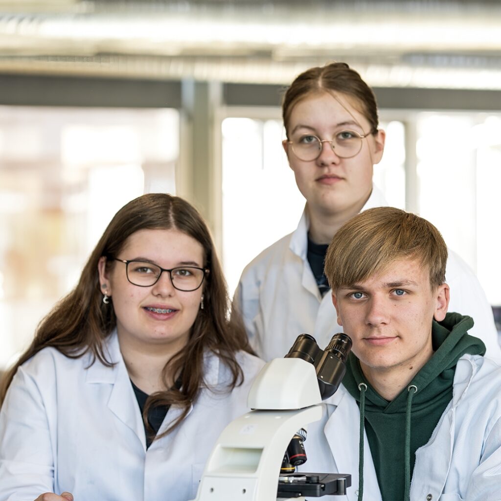 Tre elever fra NEG Kalundborg er iført kitler og står omrking et mikroskop.
