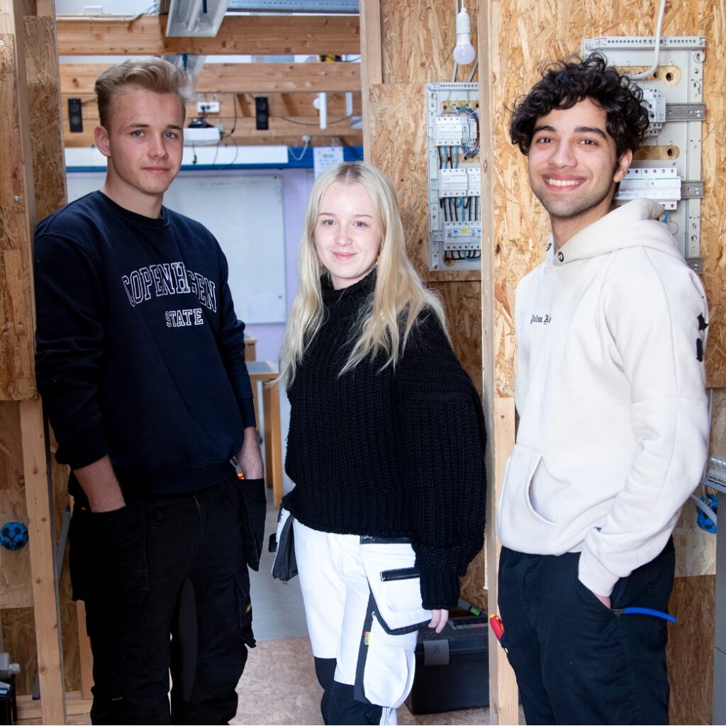 Tre elektrikerelever fra NEG Kalundborg står og smiler til kameraet. De står foran deres stande de har sat op i undervisningen.