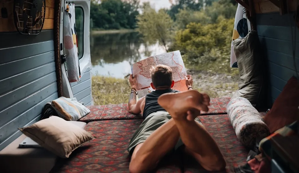 En ung fyr ligger og kigger på et kort, i en autocamper. Der er udsigt til en sø og et grønt område.